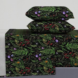 Puede incluir: Juego de cama negro con diseño botánico. El juego incluye dos almohadas y una sábana bajera, todas con un estampado de helechos verdes, flores moradas y amarillas y bayas rojas. El diseño está sobre un fondo negro.