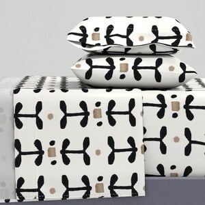 Puede incluir: Juego de sábanas blancas con un patrón abstracto repetido en negro y beige. Dos fundas de almohada están apiladas encima de las sábanas. El patrón presenta formas negras y rectángulos y círculos beige. Medidas métricas.