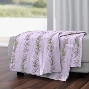 May include: A lavender throw blanket with a floral design. The pattern shows vertical rows of purple flowers and green leaves on a light purple background. The blanket is draped over a grey ottoman.