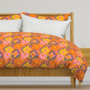 May include: A duvet cover and two pillow shams featuring a repeating pattern of orange, yellow, and pink daisies set against a background of wavy grey and orange lines. The bedding set is draped over a wooden bed frame with white pillows.