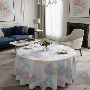 Pode incluir: Uma toalha de mesa redonda com um design em aquarela em tons pastel de azul, roxo e pêssego. O tecido é colocado sobre uma mesa redonda, com um vaso de flores no centro. A sala tem um sofá, uma cadeira e obras de arte.