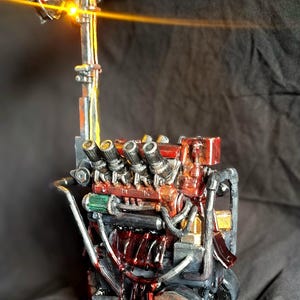 May include: A miniature mechanical engine sculpture featuring a tall vertical light pole with three circular lamps, one emitting a bright yellow glow. The engine block is painted in shades of red, silver, and black with intricate piping and wires.