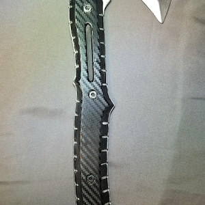Pode incluir: Um machado tomahawk prateado e preto com cabo de fibra de carbono. A cabeça do machado tem uma lâmina afiada e uma ponta. O cabo é texturizado para aderência e possui rebites de metal. Cerca de 40 cm de comprimento.
