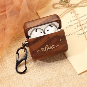 May include: A wooden AirPods case with the word "love" in cursive script. The case is open, revealing white earbuds. A black carabiner clip is attached. A handwritten note with text is in the background.