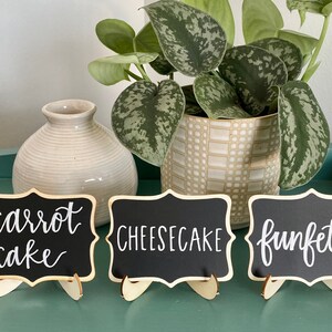 May include: Three small black chalkboard signs with white calligraphy on wooden stands. The signs read "carrot cake", "cheesecake", and "funfetti".