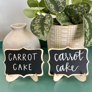 May include: Two black chalkboard signs with wooden stands, each displaying the text "Carrot Cake" in white chalk. The signs are sitting on a green surface in front of a potted plant.