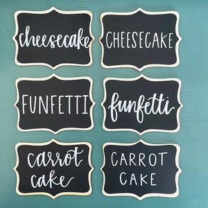 May include: Six black chalkboard signs with white calligraphy. The signs read "cheesecake", "FUNFETTI", "carrot cake", "CHEESECAKE", "funfetti", and "CARROT CAKE".
