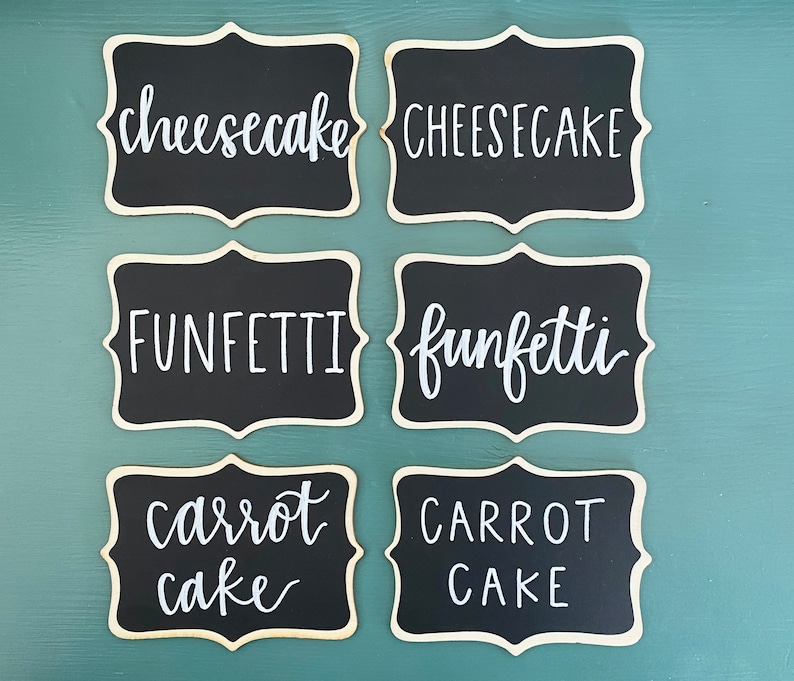 May include: Six black chalkboard signs with white calligraphy. The signs read "cheesecake", "FUNFETTI", "carrot cake", "CHEESECAKE", "funfetti", and "CARROT CAKE".