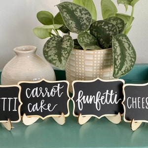 May include: Four small black chalkboard signs with white calligraphy on wooden stands. The signs read "Funfetti", "Carrot Cake", "Funfetti", and "Cheesecake".