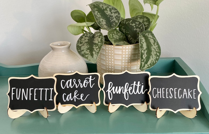 May include: Four small black chalkboard signs with white calligraphy on wooden stands. The signs read "Funfetti", "Carrot Cake", "Funfetti", and "Cheesecake".