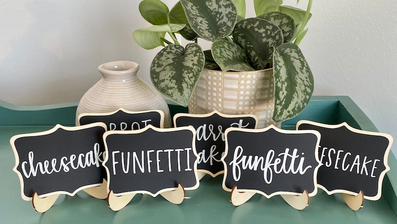 May include: Six small black chalkboard signs with white lettering on wooden stands. The signs read "cheesecake", "funfetti", "carrot cake", "funfetti", and "cheesecake".