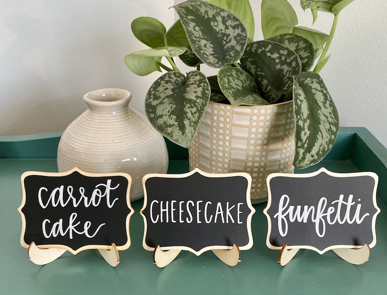 May include: Three small black chalkboard signs with white calligraphy on wooden stands. The signs read "carrot cake", "cheesecake", and "funfetti".