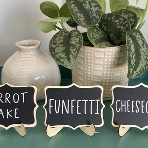 May include: Three small black chalkboard signs with white lettering on wooden stands. The signs read "Carrot Cake", "Funfetti", and "Cheesecake".