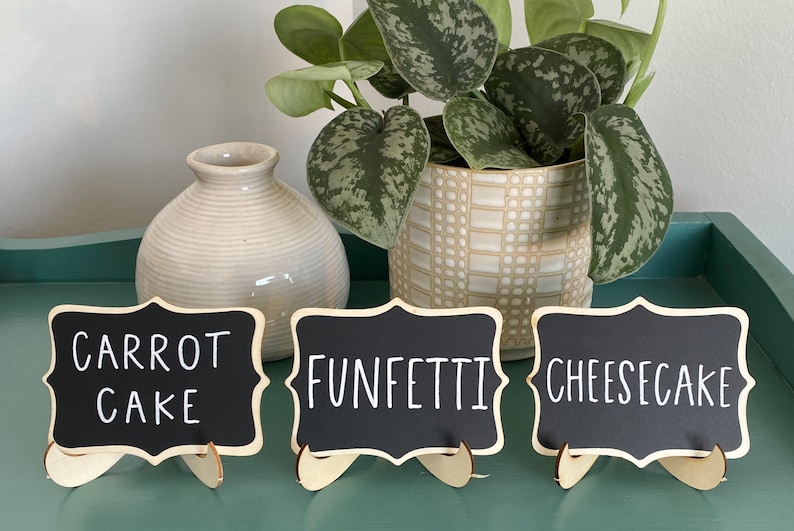May include: Three small black chalkboard signs with white lettering on wooden stands. The signs read "Carrot Cake", "Funfetti", and "Cheesecake".