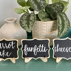 May include: Three small black chalkboard signs with white calligraphy on wooden stands. The signs read "carrot cake", "funfetti", and "cheesecake".
