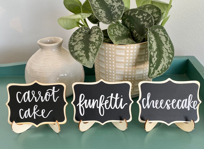 May include: Three small black chalkboard signs with white calligraphy on wooden stands. The signs read "carrot cake", "funfetti", and "cheesecake".