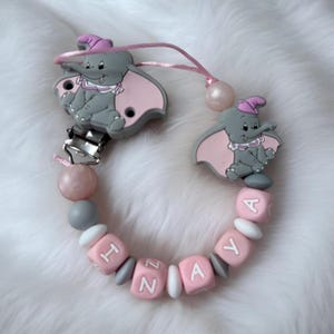Peut inclure: Un attache-tétine pour bébé avec deux breloques grises représentant Dumbo avec des oreilles et des chapeaux roses. Le clip comprend des perles roses et grises, et des blocs roses avec un nom. Le clip est attaché à un clip en métal.