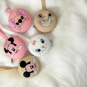 Könnte beinhalten: Eine Sammlung runder Münzbörsen mit Reißverschluss, die mit Cartoon-Charakteren verziert sind. Die Geldbörsen sind in verschiedenen Farben erhältlich, darunter Rosa, Beige und Weiß, mit Charakteren wie Stitch, Simba, Minnie Mouse, Marie und Mickey Mouse. Jede Geldbörse hat eine Handschlaufe.