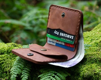 May include: A brown leather wallet with a flap closure and a snap button. The wallet is open and has several cards and cash inside. The wallet is sitting on a bed of green moss.