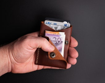 Könnte beinhalten: Ein braunes Lederportemonnaie mit Druckknopfverschluss, das mehrere Kreditkarten und einen 20-Euro-Schein enthält. Das Portemonnaie ist geöffnet und zeigt seinen Inhalt. Die Nähte sind sichtbar, und das Leder hat eine natürliche Textur.