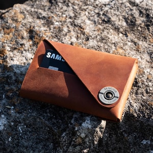 Könnte beinhalten: Eine braune Ledergeldbörse mit einem Metallknopf, auf dem "MAGOR" und "HANDCRAFTED ELEGANCE" steht. Eine schwarze Kreditkarte mit dem Wort "SAMSUNG" ist teilweise in der Geldbörse sichtbar.