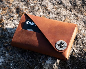 Könnte beinhalten: Eine braune Ledergeldbörse mit einem Metallknopf, auf dem "MAGOR" und "HANDCRAFTED ELEGANCE" steht. Eine schwarze Kreditkarte mit dem Wort "SAMSUNG" ist teilweise in der Geldbörse sichtbar.
