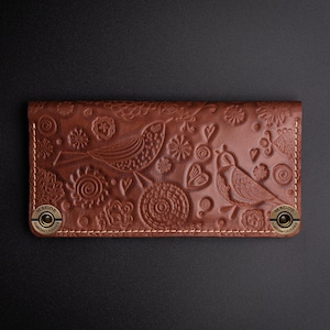May include: A brown leather wallet with a floral and bird design. The wallet has two brass snaps on the sides and the word "MAGOR" is embossed on the snaps.