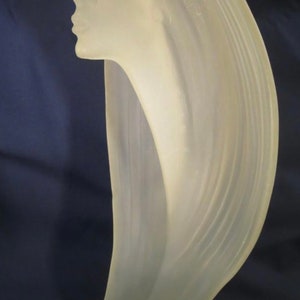 Op de afbeelding: Een doorschijnende sculptuur van een vrouwenprofiel, met vloeiend haar. De sculptuur heeft een bleke, crèmekleurige kleur en heeft een gladde, gepolijste afwerking. Het haar stroomt in een gebogen vorm naar beneden, waardoor een sierlijke en elegante vorm ontstaat. De sculptuur staat tegen een donkerblauwe achtergrond.