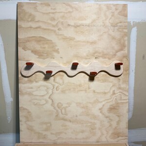 May include: A light-colored wooden wall-mounted coat rack featuring a wavy, serpentine design. The rack includes five small, reddish-brown pegs attached to the front, positioned along the curves of the wood to provide hanging points for items.