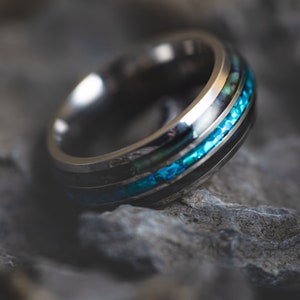 May include: A close-up of a silver ring with a black band and a vibrant turquoise inlay. The ring is set against a textured gray stone background, highlighting its intricate design and craftsmanship.