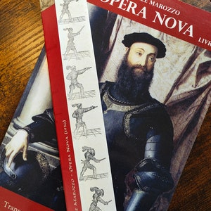 Peut inclure: Un marque-page avec des illustrations d'escrimeur sur un livre rouge intitulé "OPERA NOVA" d'Achille Marozzo. Le livre et le marque-page sont posés sur une surface en bois, créant une ambiance classique.