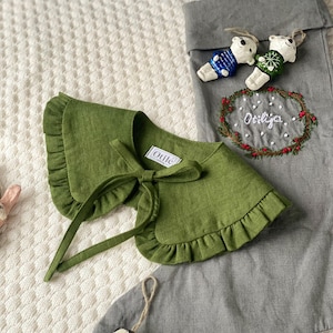 May include: A green linen collar with a ruffled edge and a bow tie, next to a gray Christmas stocking. The collar has the word "Otilé" on a tag. Two small bear ornaments and two black and white ornaments are also visible.