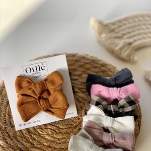 Peut inclure: Un ensemble de 9 nœuds en tissu pour cheveux dans différentes couleurs et motifs, dont le marron, le noir, le rose, le blanc et le gris. Les nœuds sont disposés sur une surface en paille tressée. Le texte "Otilé clothing" est visible sur une carte blanche.