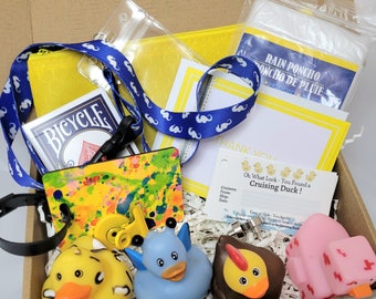 May include: A yellow box filled with a variety of items, including a deck of playing cards, a blue lanyard with a yellow and blue pattern, a yellow pouch, a clear plastic bag, a white rain poncho, a yellow and blue card with the text "Thank You", a yellow and blue card with the text "Oh What Luck - You Found a Cruising Duck!" and four rubber ducks: a yellow duck with black spots, a blue duck with a blue and white pattern, a brown duck with a yellow beak and a pink duck with a red pattern.