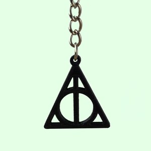 May include: Black metal keychain charm shaped like the Deathly Hallows symbol from the Harry Potter series. The symbol is a triangle with a circle inside and a vertical line through the circle.