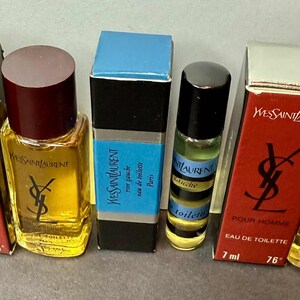 May include: Collection of Yves Saint Laurent miniature perfumes. The bottles are in various shapes and sizes, with red, black, and blue packaging. The text "POUR HOMME" and "EAU DE TOILETTE" are visible on the boxes. Each bottle contains 7 ml.