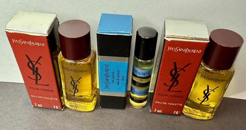 May include: Collection of Yves Saint Laurent miniature perfumes. The bottles are in various shapes and sizes, with red, black, and blue packaging. The text "POUR HOMME" and "EAU DE TOILETTE" are visible on the boxes. Each bottle contains 7 ml.