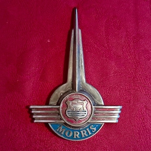 Op de afbeelding: Een vintage metalen Morris auto-embleem met een centraal rond embleem met een rode achtergrond en een zilveren stier-icoon. Het embleem heeft horizontale zilveren vleugels, een verticale vin aan de bovenkant en een blauwe boog met de tekst MORRIS.