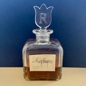 May include: A vintage perfume bottle with a clear glass tulip-shaped stopper featuring the letter "R". The rectangular bottle has a label that reads "Réplique, Raphael Paris". The liquid inside is amber-colored.