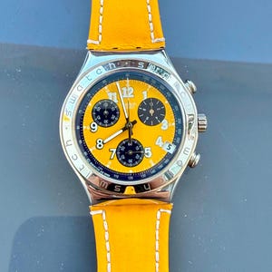 May include: A Swatch chronograph wristwatch featuring a bright yellow dial with black sub-dials, white Arabic numerals, and a date window at the four o'clock position. The watch has a silver-tone metal case and a vibrant yellow leather strap.