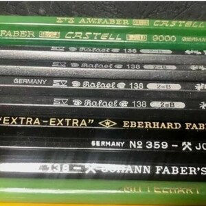 May include: Close-up of a collection of black and green pencils with various markings, including "EXTRA-EXTRA" Eberhard Faber 1040, "Germany No. 359 - Johann Faber No. 318", and "A.W. Faber Castell 9000 Germany".