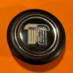 Könnte beinhalten: Ein schwarzes, kreisförmiges Emblem mit silbernem Rand. Das Emblem zeigt ein stilisiertes, weißes und blaues Design in der Mitte. Der Hintergrund ist leuchtend orange.