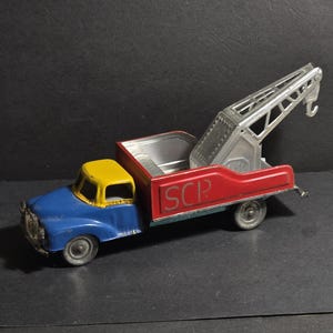 May include: A vintage toy tow truck with a blue cab, yellow roof, red bed, and a silver crane. The bed has the letters "SCR" printed on it.