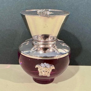 Può includere: Un flacone di vetro viola con un tappo argentato e un design a testa di Medusa argentato. Il flacone è riempito con un liquido trasparente.