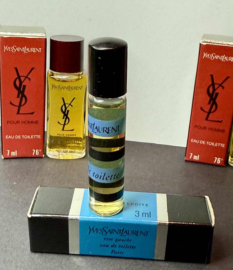 May include: A collection of Yves Saint Laurent miniature perfume bottles and boxes. The bottles are cylindrical with black caps and striped labels. The boxes are red and black with the Yves Saint Laurent logo and product information.