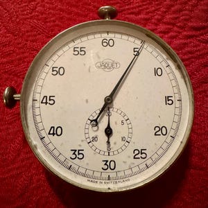 Op de afbeelding: Een vintage zilveren Jaquet stopwatch met een witte wijzerplaat en zwarte wijzers. De stopwatch heeft een grote buitenste wijzerplaat met nummers van 5 tot 60 en een kleinere binnenste wijzerplaat met nummers van 5 tot 30. De stopwatch is gemaakt in Zwitserland.