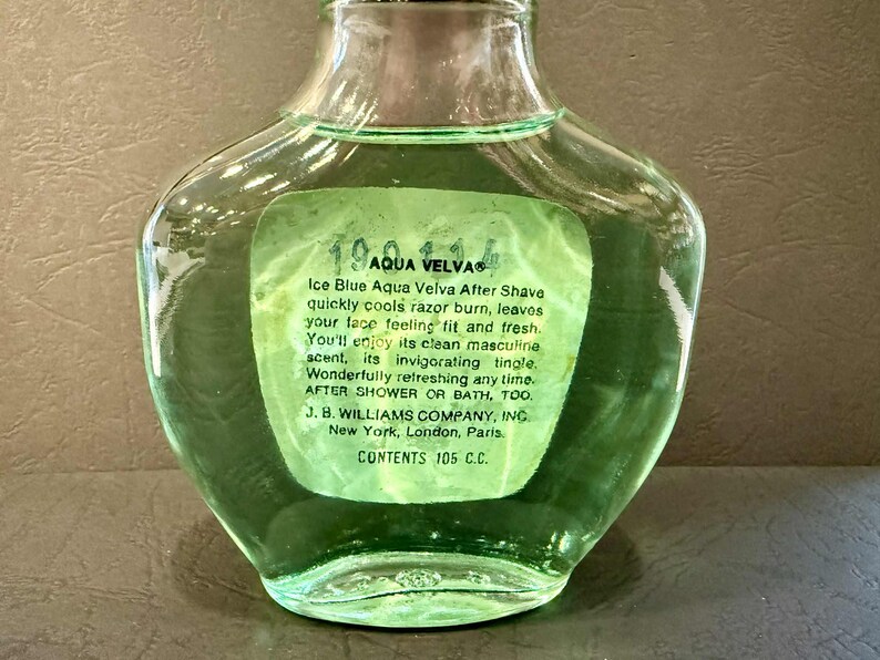 Puede incluir: Una botella de vidrio transparente de loci&oacute;n para despu&eacute;s del afeitado Aqua Velva con una etiqueta verde. La etiqueta incluye el texto "Ice Blue Aqua Velva After Shave" y otros detalles del producto. La botella contiene 105 ml.