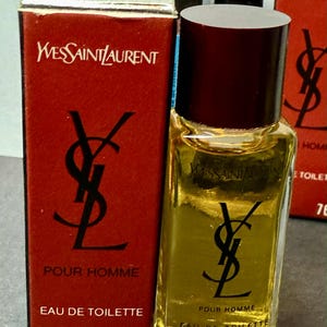 May include: A small bottle of Yves Saint Laurent Pour Homme Eau de Toilette, with a dark burgundy cap and a clear glass body. The bottle is next to a red box with the YSL logo and text. The bottle contains 7 ml.