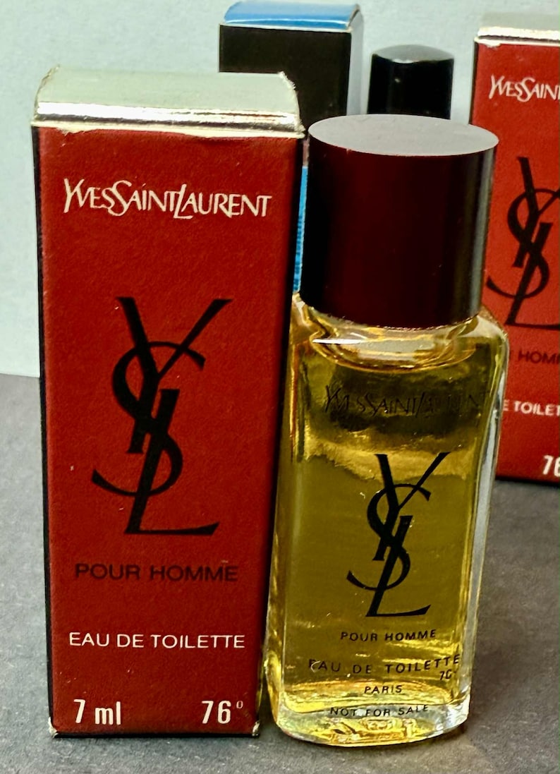 May include: A small bottle of Yves Saint Laurent Pour Homme Eau de Toilette, with a dark burgundy cap and a clear glass body. The bottle is next to a red box with the YSL logo and text. The bottle contains 7 ml.