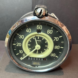 May include: A vintage chrome speedometer with a black face and white numbers. The speedometer reads up to 90 and has a smaller clock face in the centre. The clock face is white with black hands and black numbers. The speedometer has a chrome bezel and a black knob on the top.